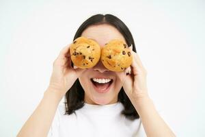 Close up portrait of funny asian girl, holds cupcakes near face, makes happy faces, white studio background photo