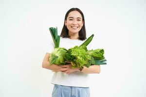 imagen de sonriente asiático mujer, vegetariano con un montón de verde verduras, comiendo oránico alimento, aislado en blanco antecedentes foto