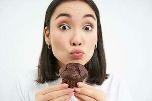 cerca arriba de linda sonriente asiático mujer, participación chocolate magdalena cerca boca, teniendo morder, disfruta comiendo Pastelería, blanco antecedentes foto