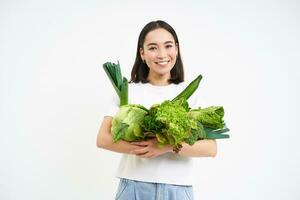 imagen de sonriente asiático mujer, vegetariano con un montón de verde verduras, comiendo oránico alimento, aislado en blanco antecedentes foto