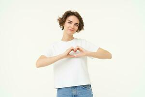 Portrait of brunette girl shows heart sign, love gesture, express care and warm feelings, stands over white background photo