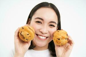 Image of korean woman enjoys eating baked pastry, showing two tasty cupcakes near face and smiles, white background photo