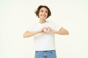 Portrait of brunette girl shows heart sign, love gesture, express care and warm feelings, stands over white background photo