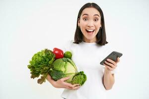 Excited korean woman, holding vegetables and smartphone, smiling and looking with joy, order organic food online, white background photo