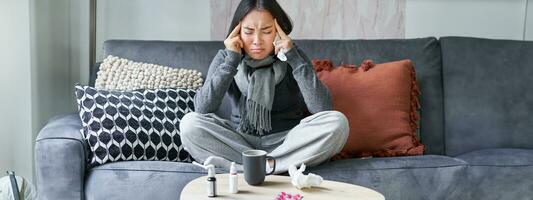 Young asian woman feels unwell, touches head, has headache and fever, staying at home on sick leave, drinking hot tea and taking drugs prescribed by GP photo