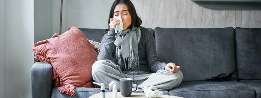 Portrait of korean woman sneezing, feeling sick, staying at home with flu or cold, neck wrapped with scarf, staying at home, taking medication photo