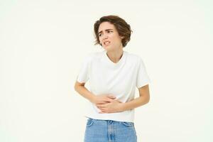 Woman grimacing from pain in stomach, stomachache, touches her belly, has painful periods, menstrual cramps, asks for painkillers, stands over white background photo