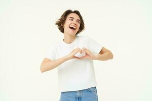 Portrait of carefree and happy woman, laughing, showing heart gesture near chest, expresses love and care, isolated over white background photo