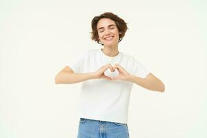 Portrait of young woman shows heart, love and care gesture, express sympathy, like something or someone, smiling happily, standing over white background photo
