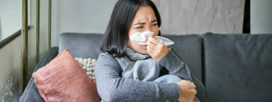 Close up of korean girl sneezing in napking, sitting at home in scarf, feeling unwell, concept of winter, heating cost and people photo