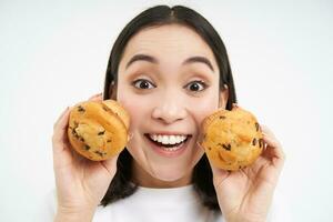 Close up portrait of asian woman, shows two cupcakes near face and smiles, likes pastry, enjoys eating bakery, white background photo