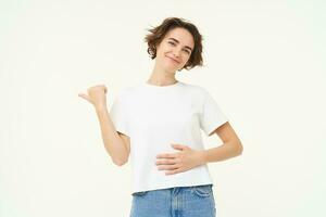 retrato de sonriente, satisfecho morena mujer, toques barriga y puntos izquierda, muestra bandera, recomienda analgésico, satisfecho por medicación efecto en su estómago, soportes terminado blanco antecedentes foto