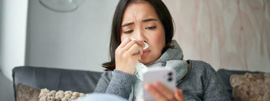 Ill korean woman sitting at home, sneezing, caught cold, reading news on mobile phone, freezing at her home because of heating problems photo