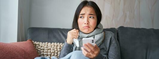 Asian girl with influenza, sits at home, cries and looks upset, holds smartphone, feels unwell and gloomy, wearing warm clothes photo