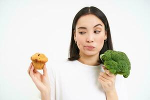 retrato de asiático niña pensando qué a comer, elegir Entre magdalena y brócoli verduras, blanco antecedentes foto