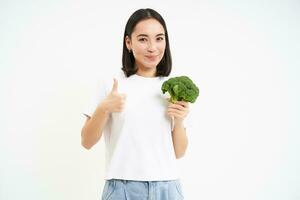 contento y sano joven mujer, muestra pulgares arriba y brócoli, delicioso vegetales para bueno dieta, blanco antecedentes foto