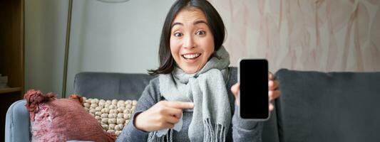 Excited young woman pointing finger at smartphone, showing online doctor, medical application or GP contact on mobile phone, staying at home sick, catching cold photo