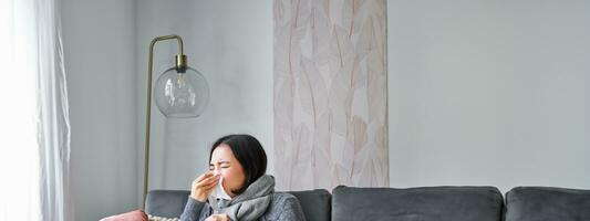 Woman with temperature, coughing, showing symptoms of flu, influenza or cold, sitting in warm clothes on sofa, covered with blanket, sneezing, staying at home in living room photo