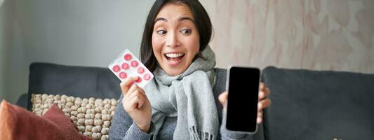 Happy asian woman catching cold, showing smartphone screen and pills, recommending online doctor app, GP or medical clinic on phone, holding drugs, staying at home with flu photo