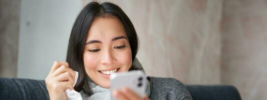 Portrait of korean woman feels sick, holding smartphone, calling doctor gp to get presctiption, caught cold, staying at home, using mobile phone photo