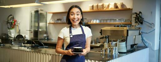 retrato de sonriente asiático hembra barista, haciendo café, participación taza de té y tomando eso a café cliente, vistiendo delantal, en pie cerca mostrador foto