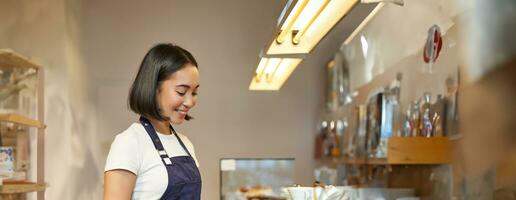 vertical Disparo de sonriente asiático niña barista, vistiendo uniforme, haciendo café, en pie cerca mostrador con taza, trabajando en café foto