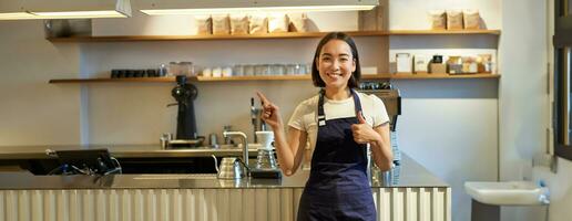 contento joven asiático barista, hembra personal trabajador en delantal, demostración pulgares arriba y señalando a Copiar espacio, demostrando café anuncio foto