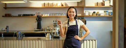 retrato de asiático niña en delantal, en pie en café cerca encimera, barista señalando dedo a Copiar espacio, vacío bandera café tienda foto