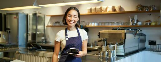 retrato de asiático sonriente hembra barista, vistiendo café delantal y participación taza de café, tomando cliente orden a el mesa, servicio bebidas foto