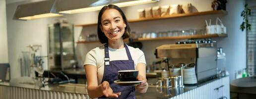 retrato de sonriente asiático mujer sostiene taza de café, preparar bebidas para clientela en cafetería, trabajando y servicio bebidas, vistiendo uniforme delantal foto