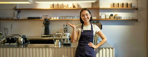 retrato de asiático niña en delantal, en pie en café cerca encimera, barista señalando dedo a Copiar espacio, vacío bandera café tienda foto