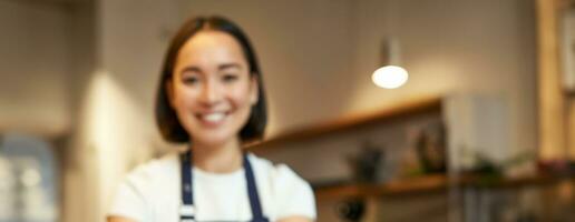 Smiling asian barista girl, giving takeaway coffee cup, prepare takeout order to guest in cafe, wearing apron uniform photo