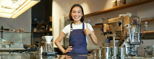 sonriente asiático barista, niña en café uniforme, muestra bueno firmar, recomienda su café, aprobar nuevo sabores, en pie en delantal foto