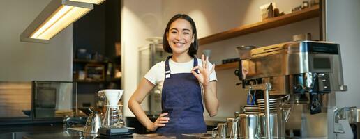 retrato de sonriente coreano barista, niña a el encimera, usa azul delantal, trabajos en café comercio, muestra bueno firmar foto