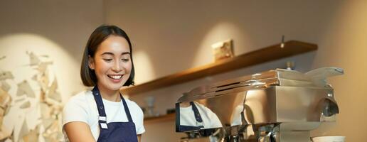 retrato de linda barista niña trabajando detrás encimera, haciendo café, humeante Leche para capuchino, vistiendo café uniforme azul delantal foto