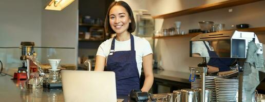 Portrait of young girl cafe owner, looking at her laptop, taking order, serving customer in coffee shop photo