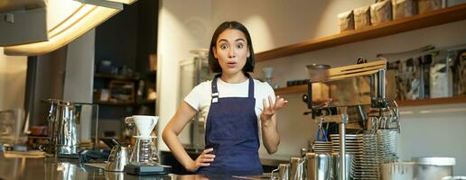 retrato de niña barista en delantal, mira sorprendido, puntos con mano y miradas asombrado a cámara, trabajos detrás mostrador y brebajes café foto