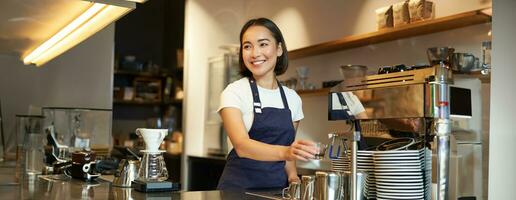 sonriente niña barista en cafetería, preparando capuchino en café máquina, humeante leche, vistiendo uniforme delantal foto