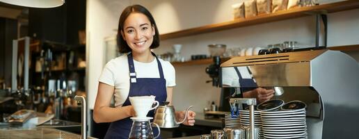 sonriente niña barista, asiático barman torrencial agua desde pava, fabricación de cerveza filtrar café en café detrás encimera, vistiendo azul delantal foto