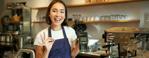 Smiling asian barista girl, wears apron, shows credit card machine for processing payment, suggest to pay contactless, standing in coffee shop photo