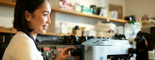 Happy asian woman, barista using coffee machine to make order, steaming milk for cappuccino and latte, laughing and smiling while working in cafe photo