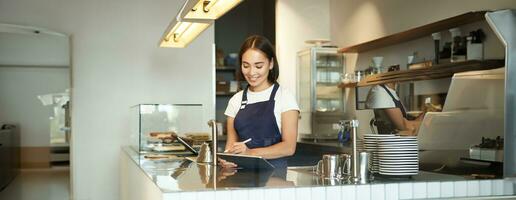 Portrait of beautiful asian girl smiling, barista in cafe working behind counter, using tablet as POS terminal, processing order photo