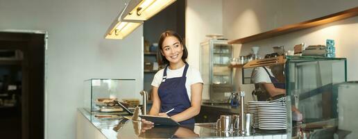 Portrait of beautiful asian girl smiling, barista in cafe working behind counter, using tablet as POS terminal, processing order photo