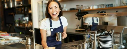 hermosa joven hembra barista, haciendo capuchino, torrencial al vapor Leche para latté Arte dentro taza, dar tu orden detrás mostrador foto