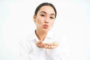 Close up portrait of cute office lady sends air kiss on palms, puckers lips and looks tender at camera, stands over white background photo