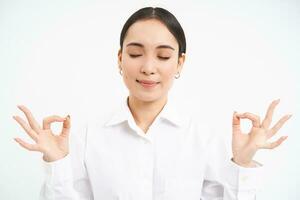 Asian business woman meditates, keeps patience, calms down, breathes in and out, relaxes after stressful day at work, stands over white background photo