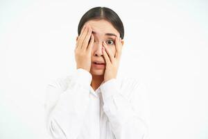 Japanese woman shuts face with hands, peeks through fingers with scared, terrified face, stands over white background photo