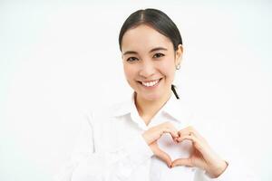 retrato de encantador coreano mujer, oficina gerente, muestra corazón mano firmar y sonriente, soportes terminado blanco antecedentes foto