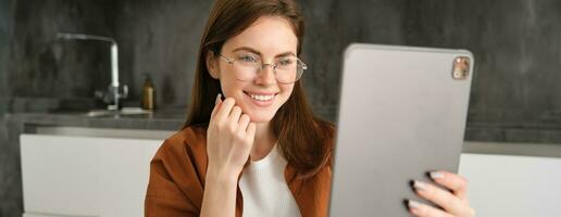 Portrait of beautiful young woman in glasses, looking at digital tablet. Businesswoman works on remote, freelancer reading on gadget, sitting in kitchen photo