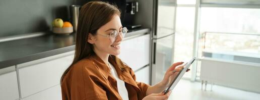 Side view of young woman in glasses, beautiful girl reading on digital tablet, messaging, online shopping with gadget, smiling, sitting in kitchen at home photo